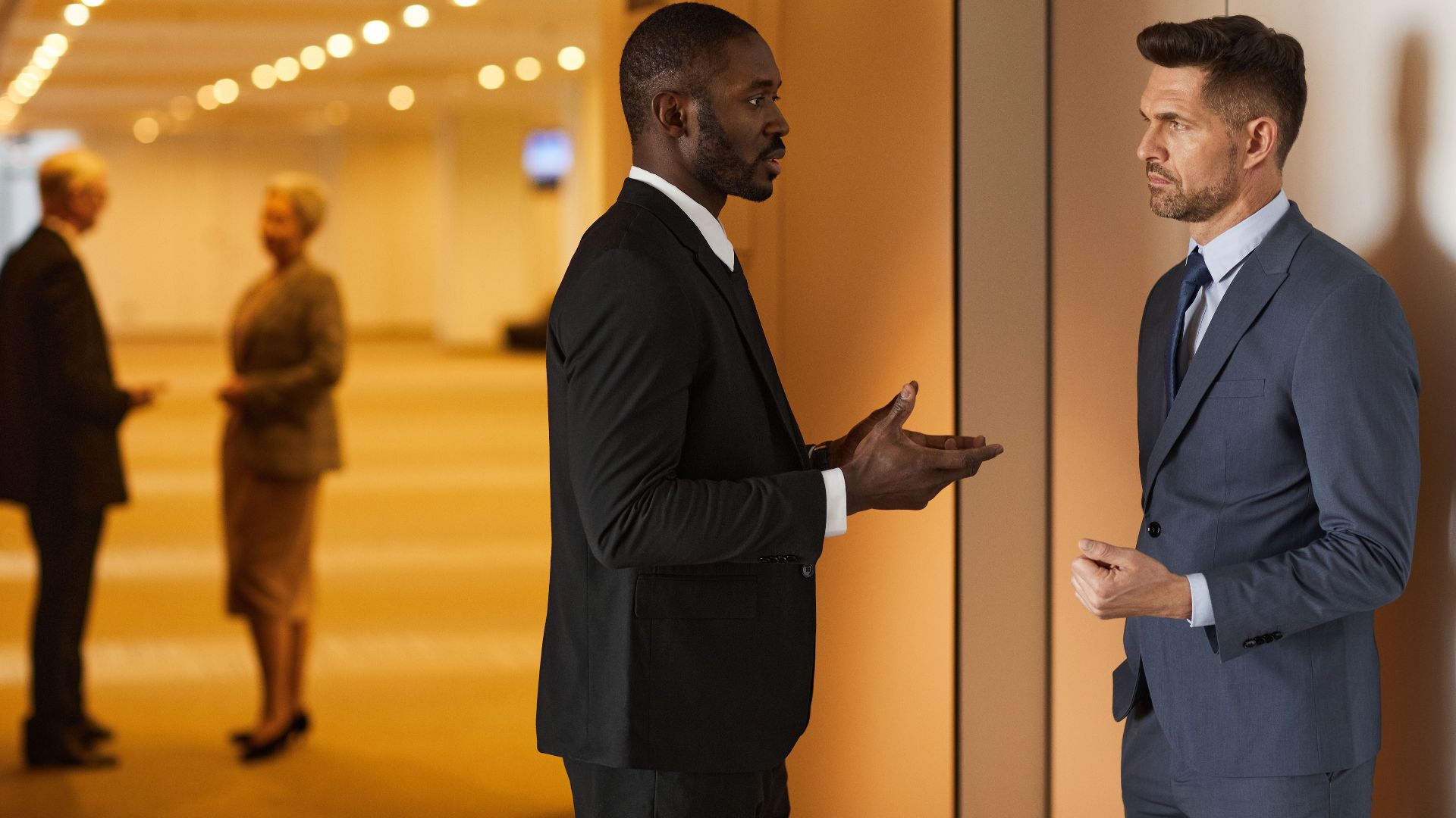 Two men in suits having a discussion in a modern hallway setting.