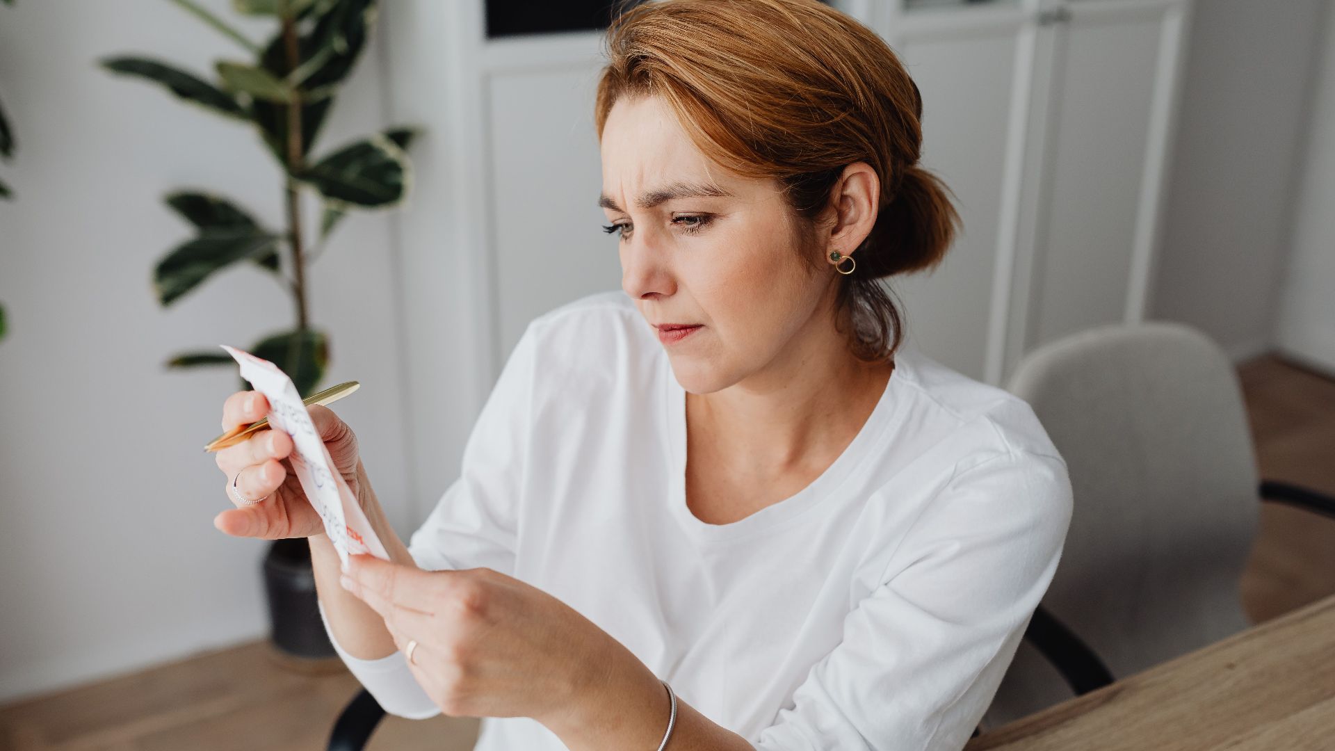 A concerned adult woman examines a receipt while sitting at her office desk, indicating financial scrutiny.