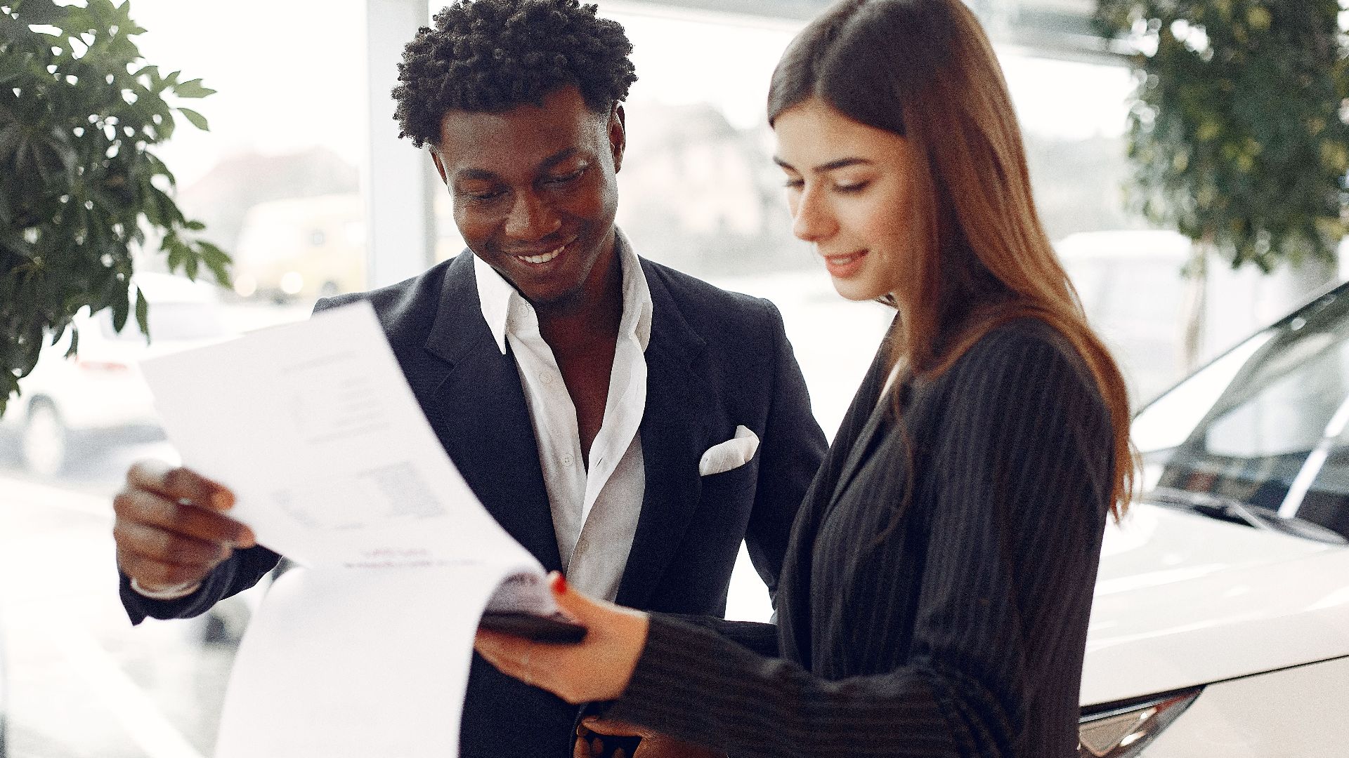 Cheerful smiling multiethnic businessman in classy suit and professional elegant female consultant standing close and reading contract details in car showroom