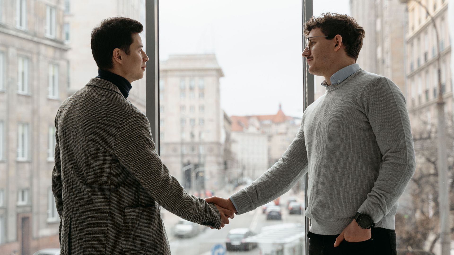 Two businessmen shaking hands in an urban office with city view through large windows.