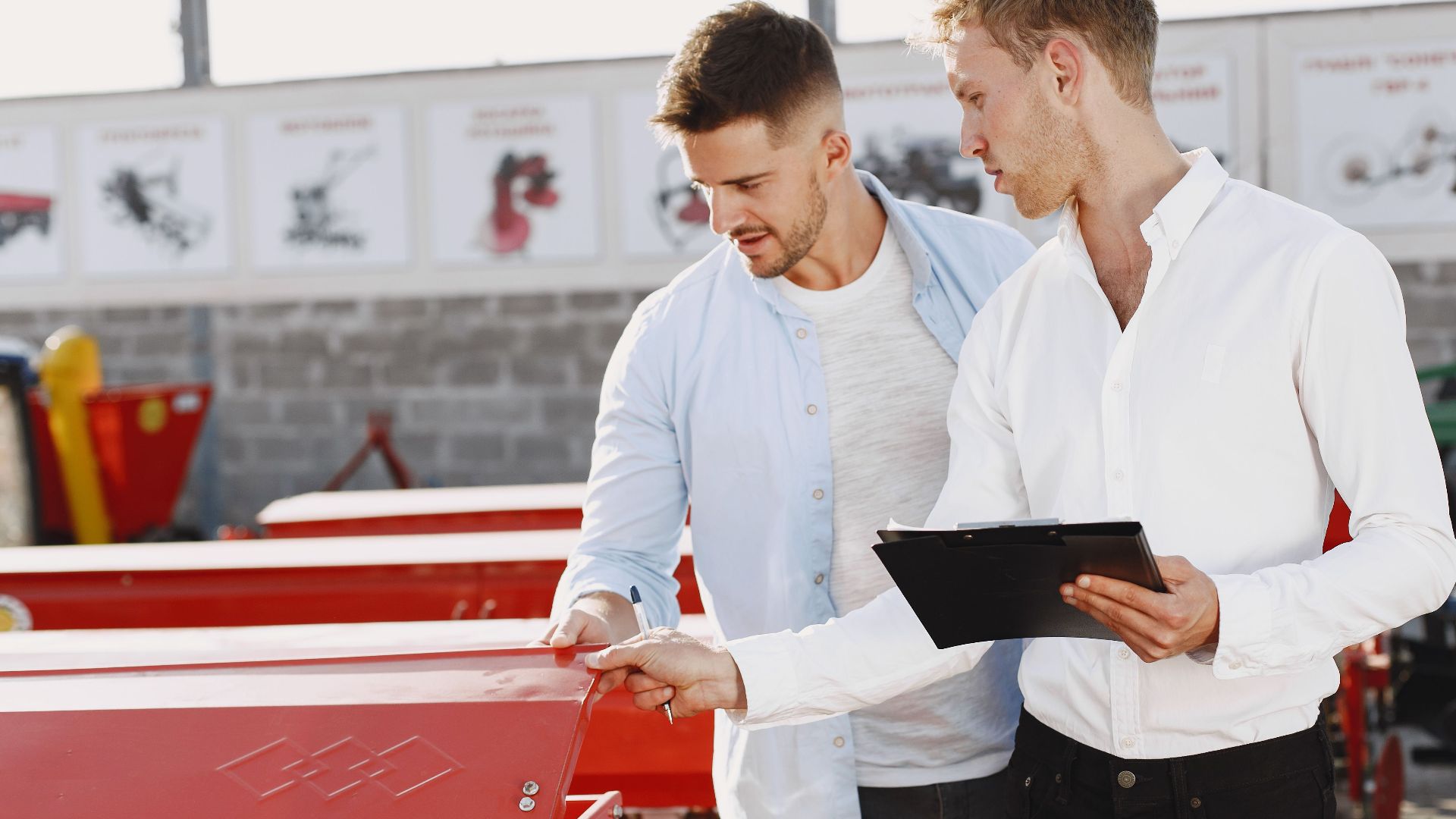Two men discussing agricultural equipment purchase at a dealership, one holding a clipboard outside.