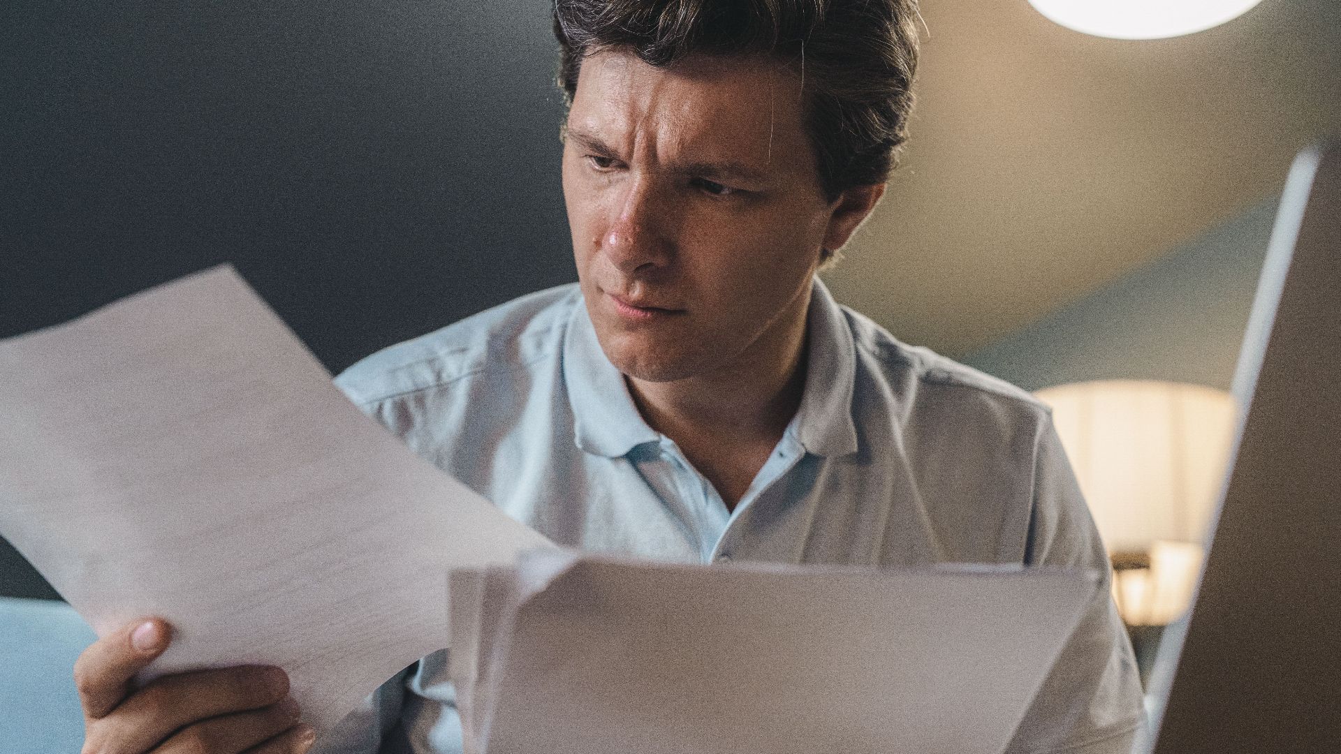 A man intently examines papers, seated indoors under warm lighting, focusing on his work.