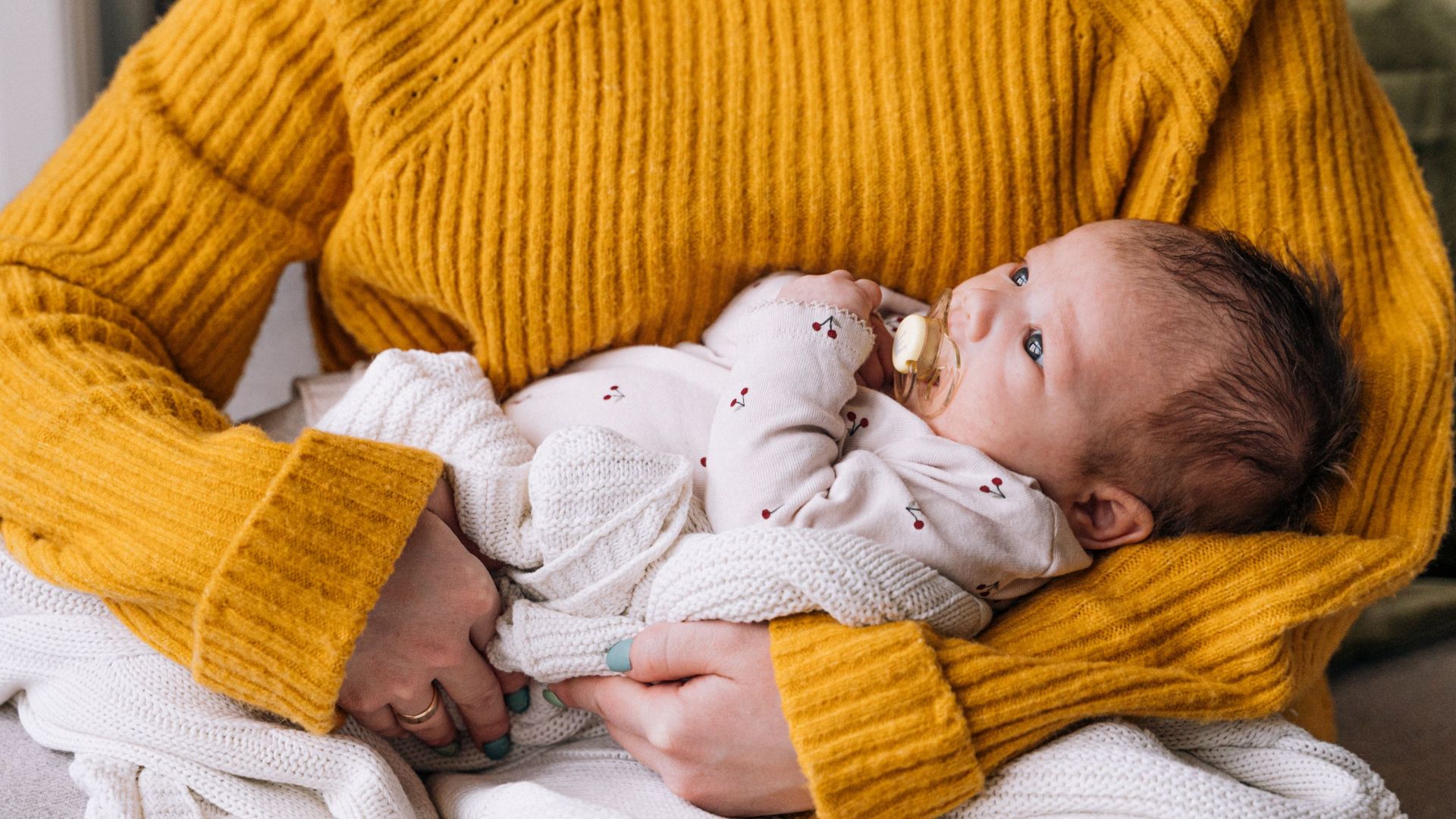 Close-up of a mother holding her newborn baby in a cozy yellow sweater indoors.