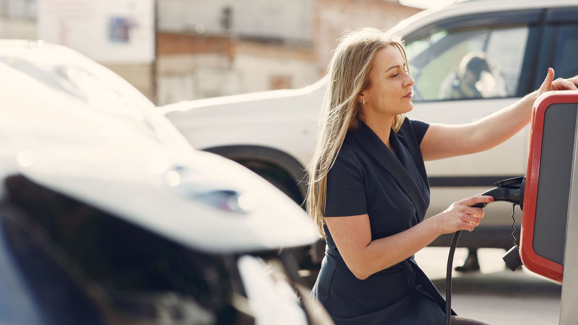 Side view of full length female driver in casual dress squatting near station while charging electric automobile for continuation of driving