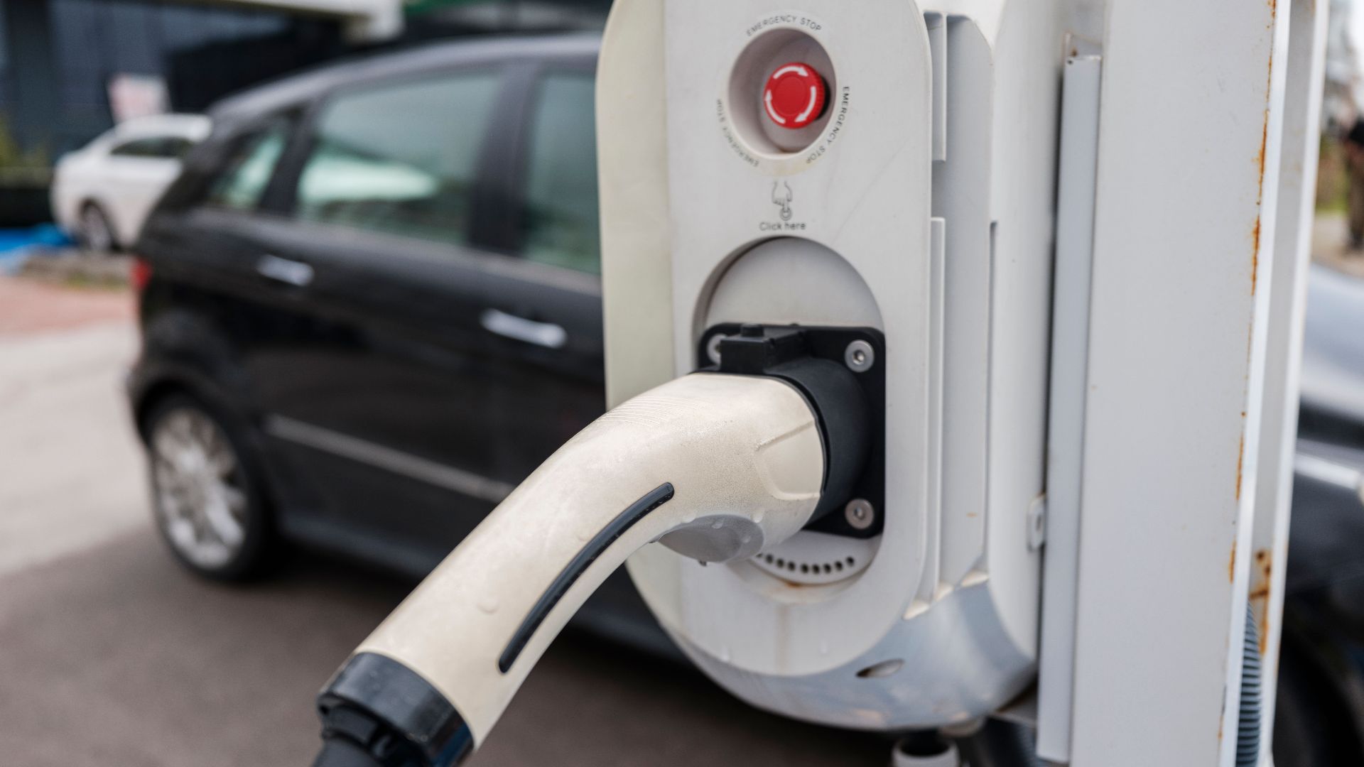 Close-up view of an electric car charging station with a plugged-in charger and cars in an urban environment.