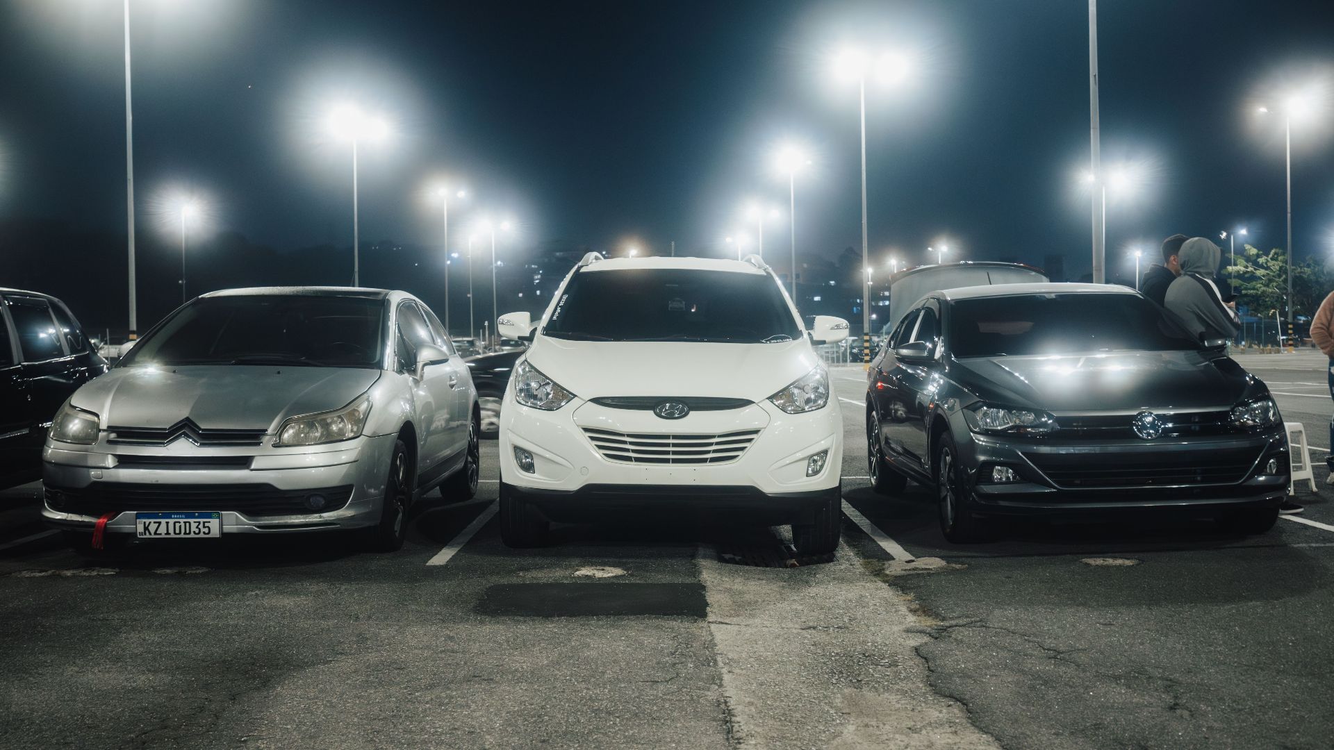 Night scene in a well-lit parking lot featuring various parked cars and street lamps.