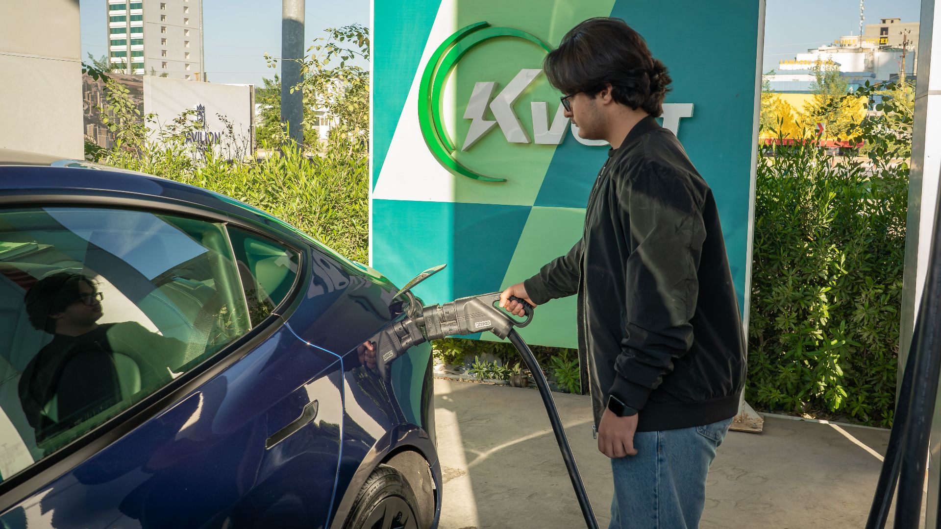 Young man charging a Tesla Model 3 at an outdoor electric vehicle station during the day.