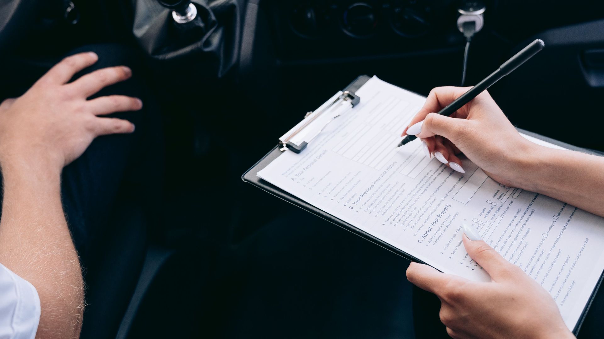 Close-up of a person writing on a clipboard inside a car, showing hands and a gear shift.