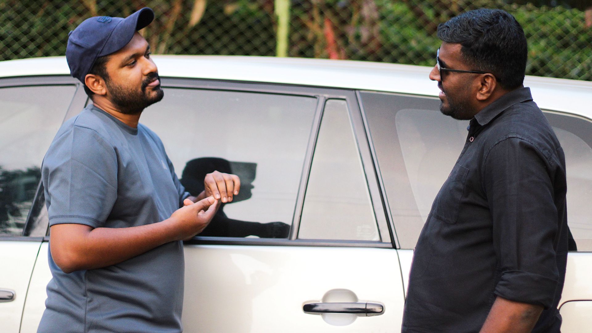 Two men having a casual discussion beside a car in an outdoor setting.