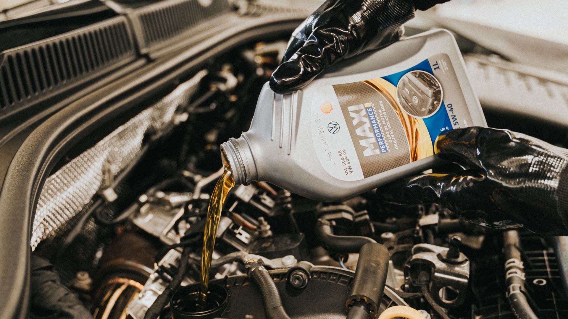 A mechanic wearing gloves pouring oil into a car engine in a garage setting.