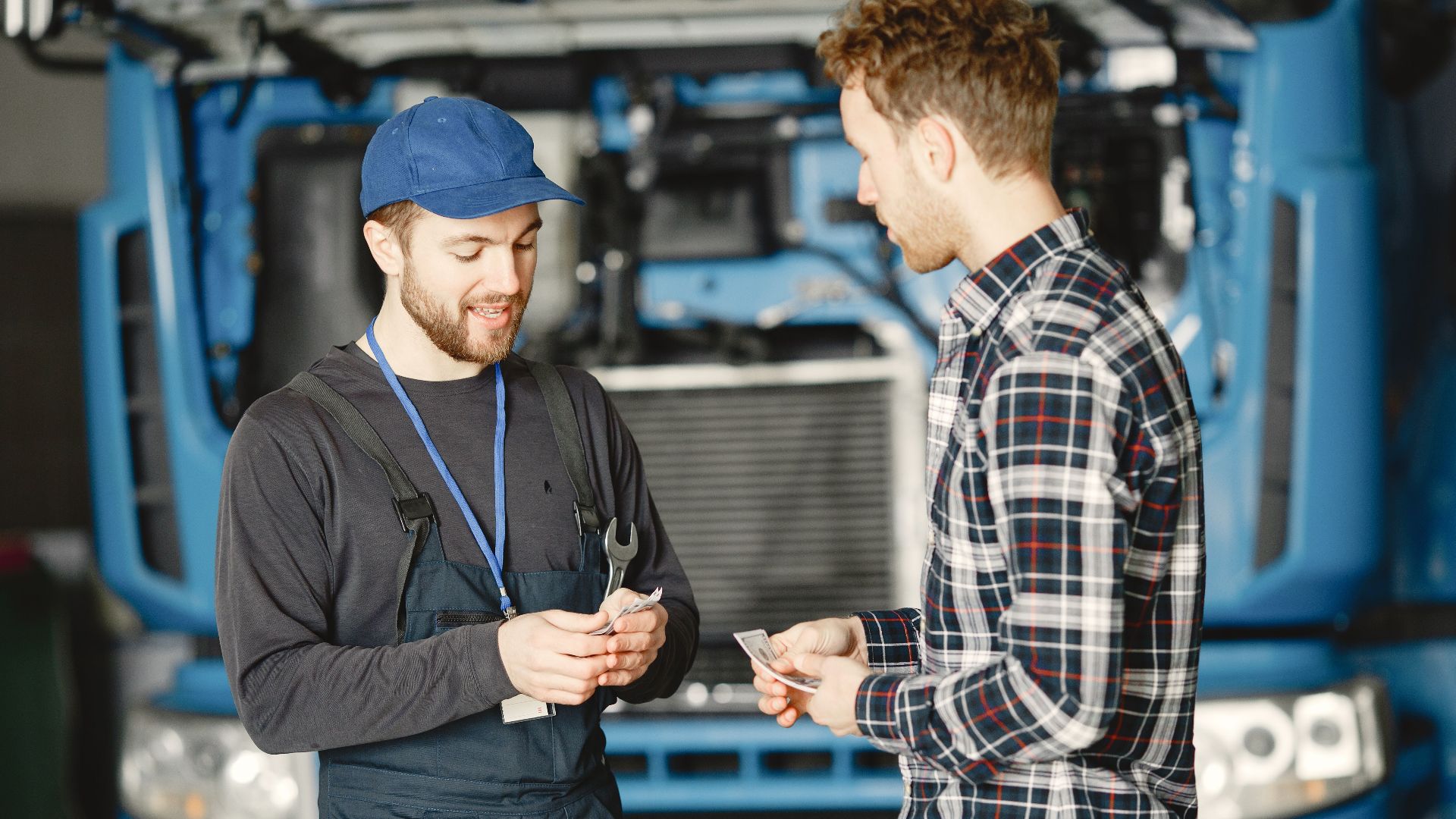 Auto mechanic in blue uniform receives payment from customer in a garage setting with a truck in the background.