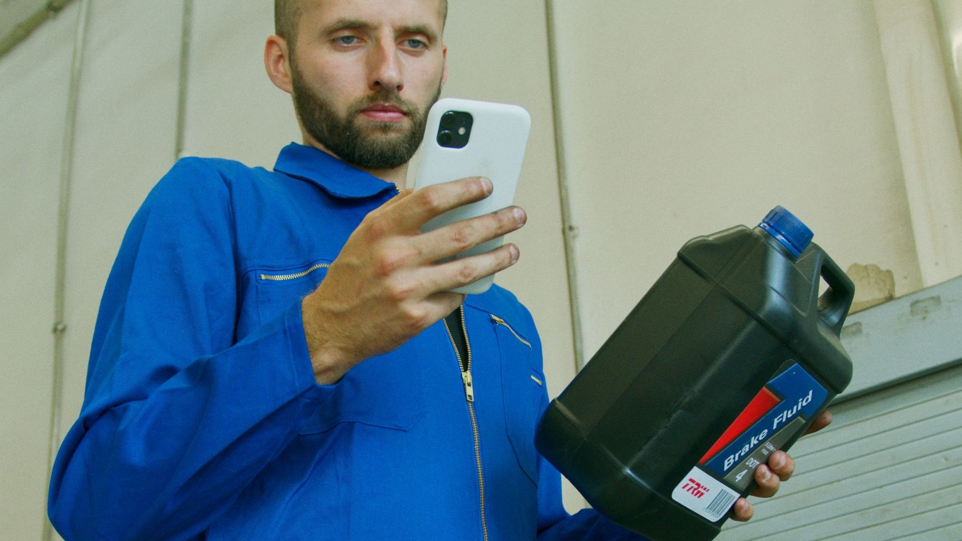 Mechanic in blue coveralls checking brake fluid container while using a smartphone in a garage workshop.