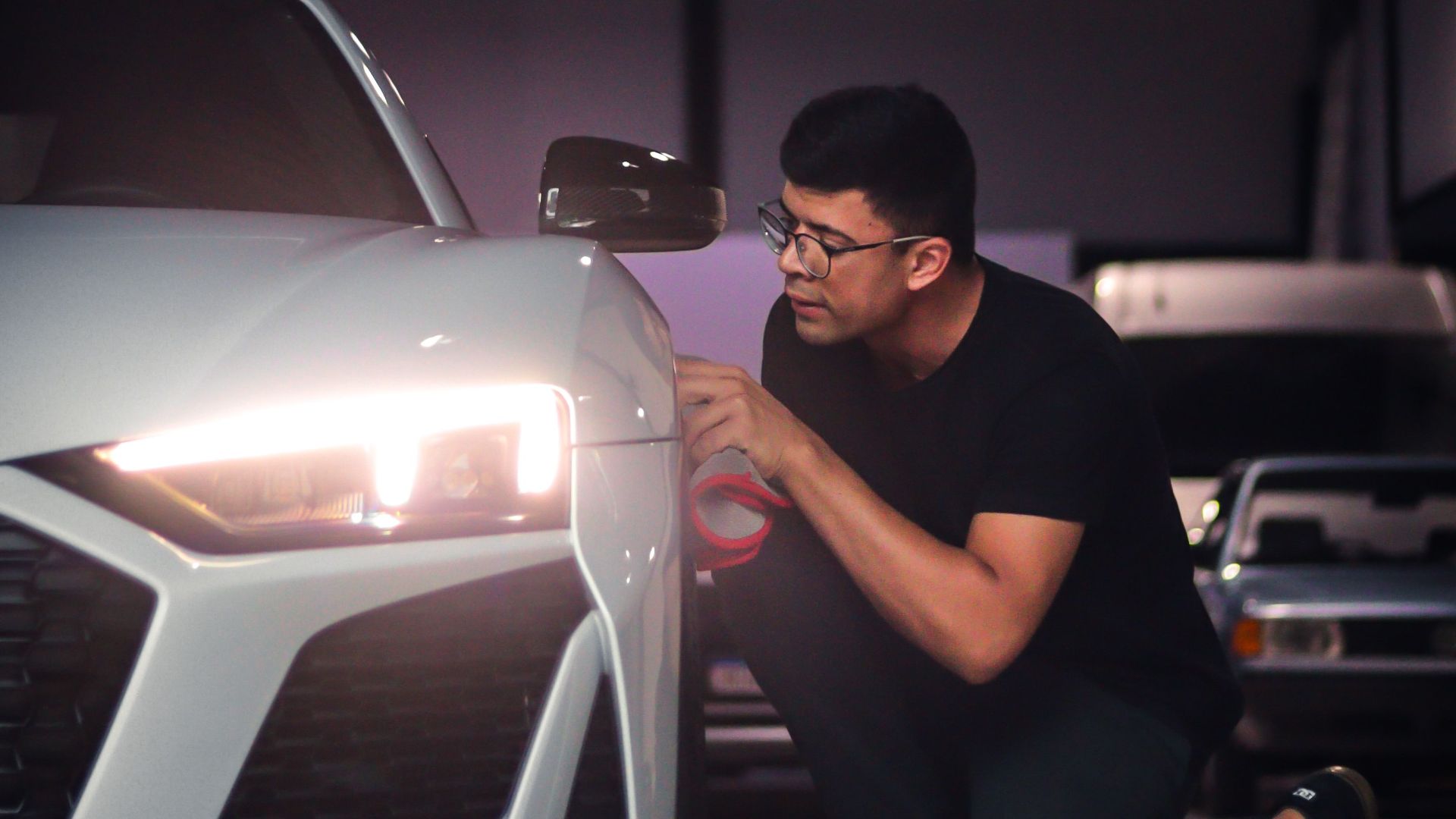 Man kneeling beside a white sports car, inspecting the headlight in a garage.