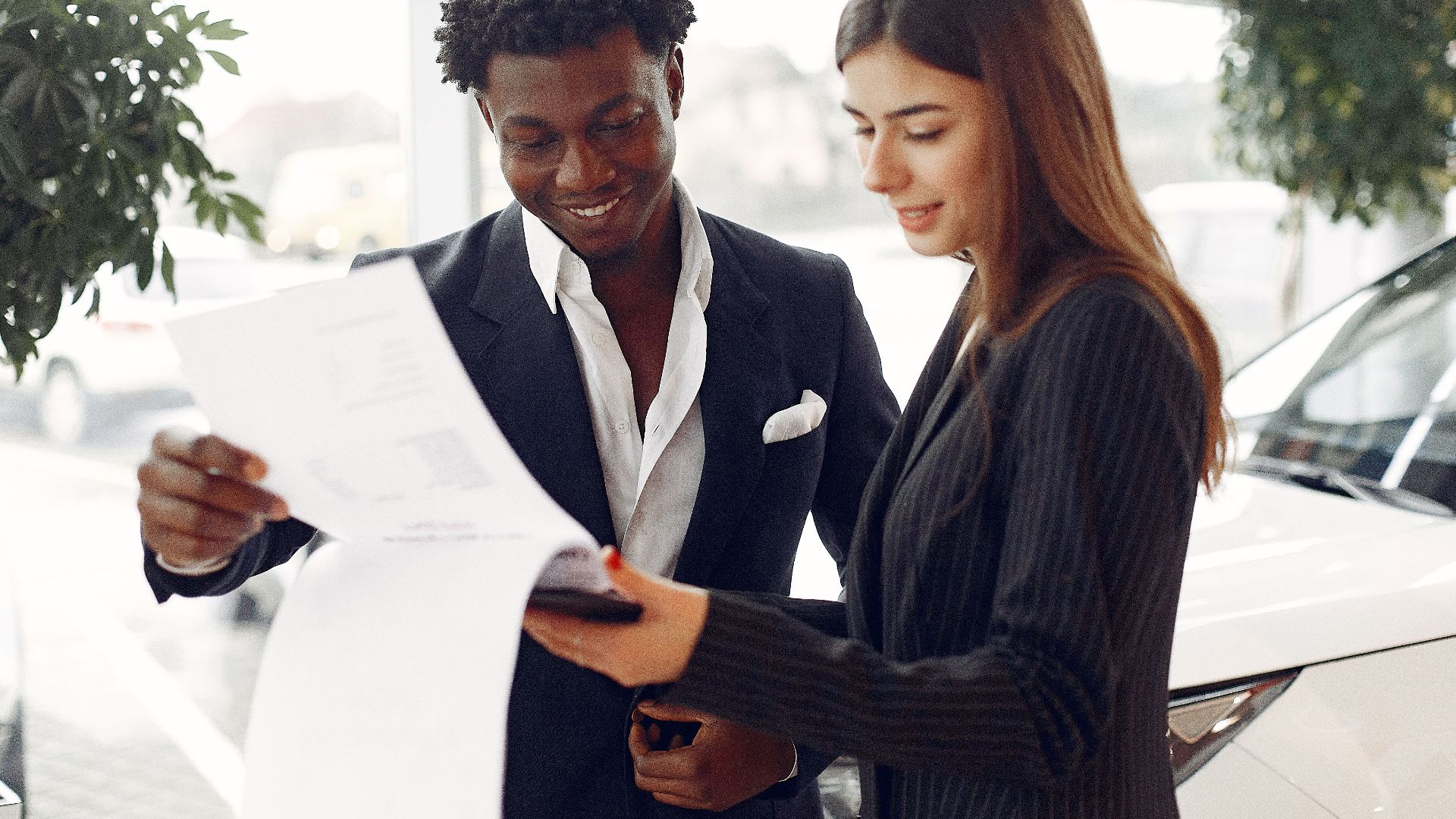 Cheerful smiling multiethnic businessman in classy suit and professional elegant female consultant standing close and reading contract details in car showroom
