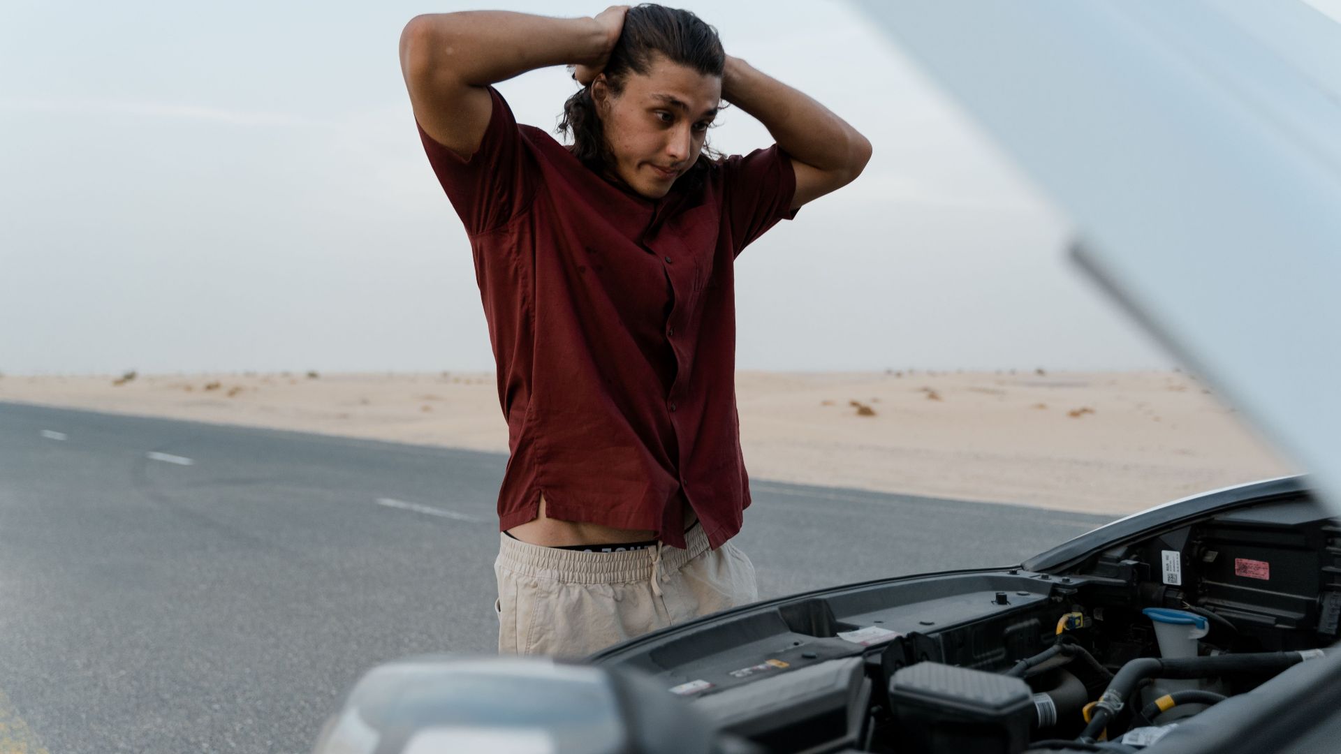 A frustrated man examining a broken car engine on a deserted road, showing stress and concern.