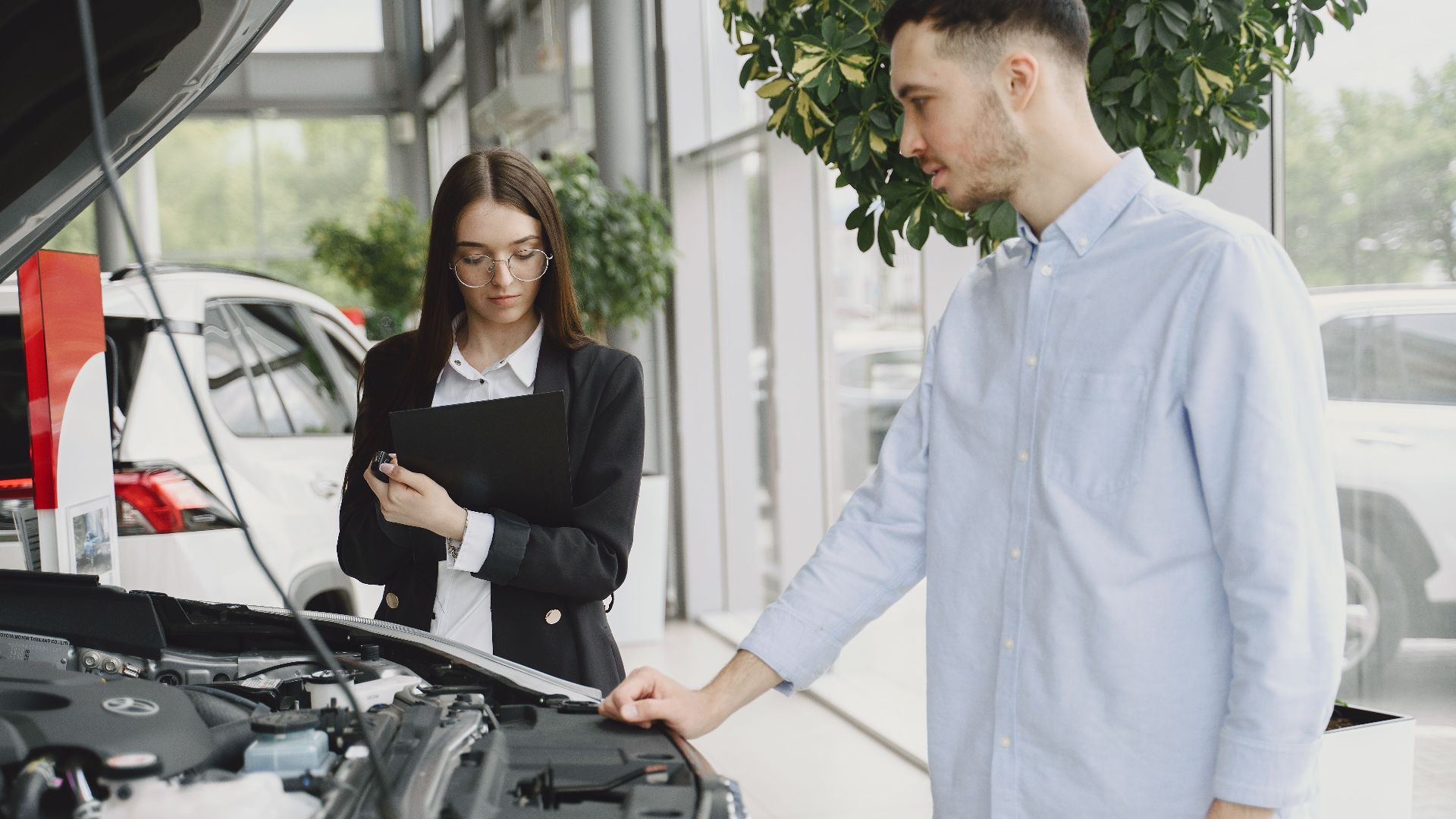 Man and woman examining car engine in dealership service area, focused on vehicle maintenance and functionality.