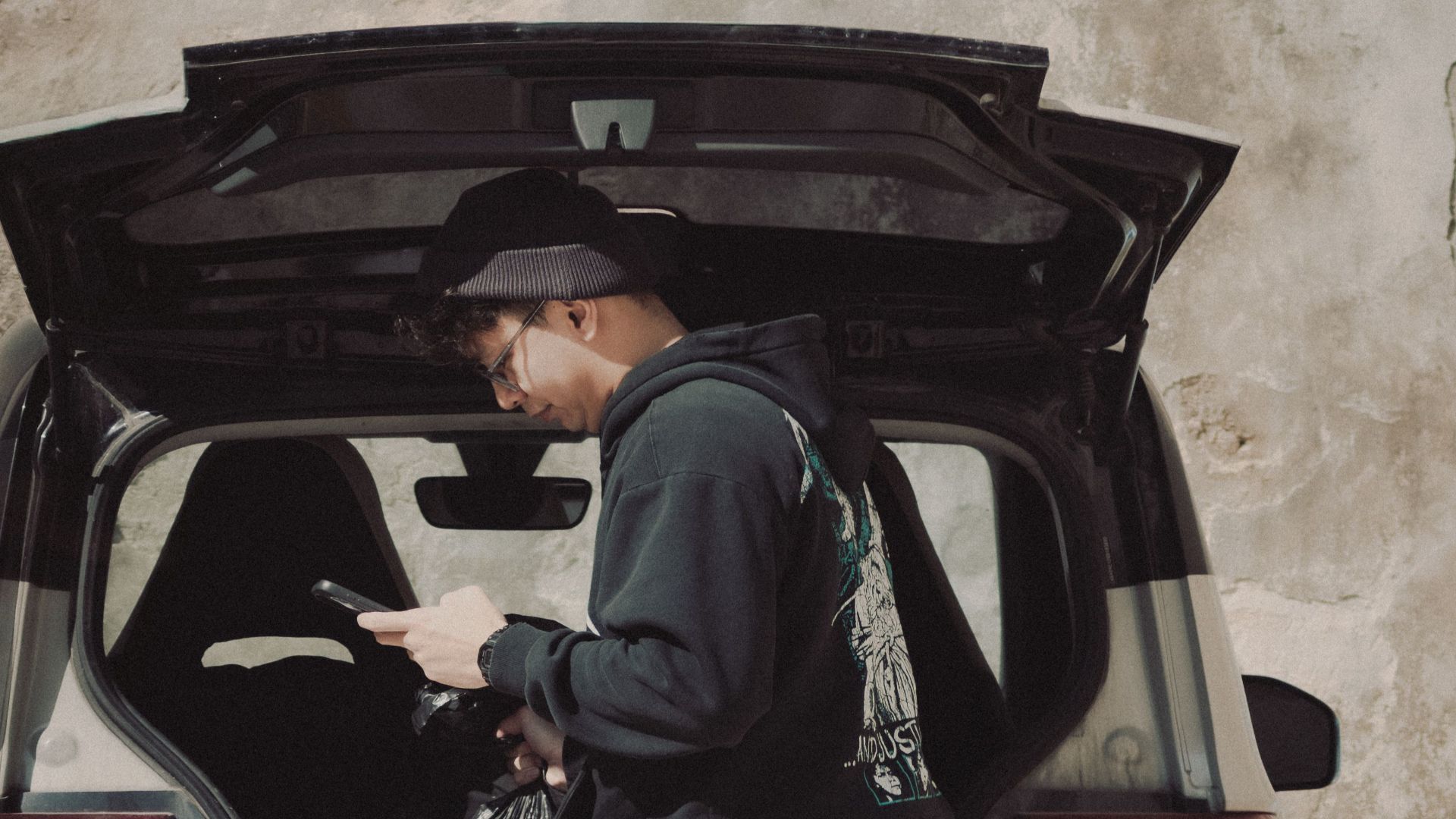 A man looks at his phone while standing by an open car trunk, outdoors.