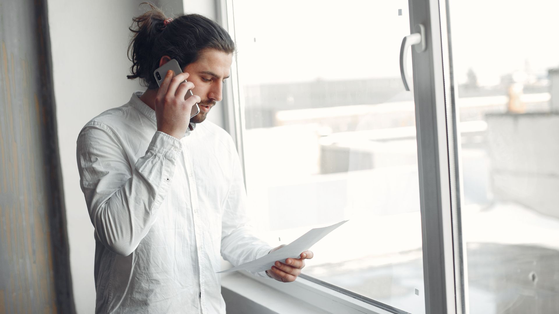 Young man in white shirt, on phone call holding a document, standing by a large window.