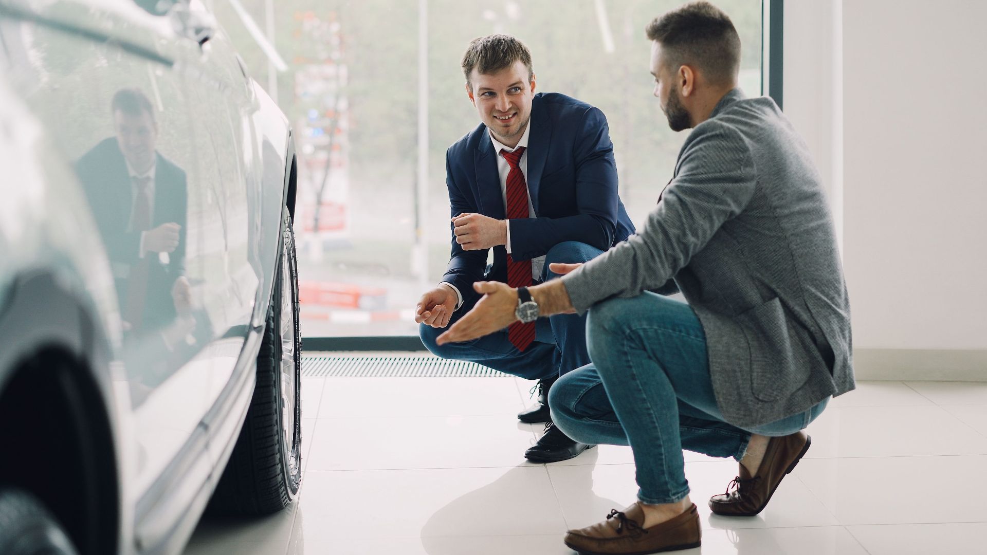 Two men discussing car features in a showroom, kneeling near a vehicle.