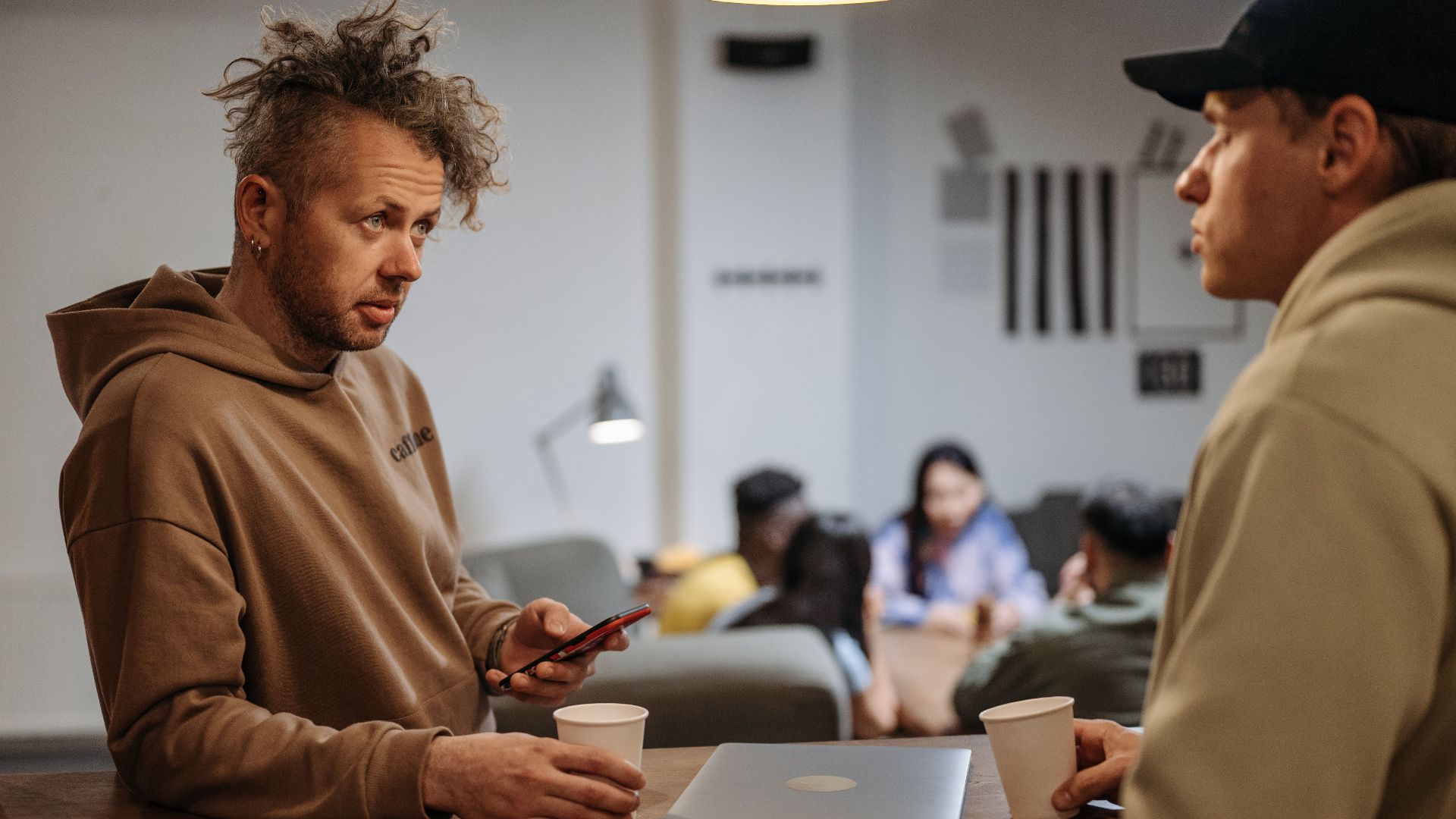 Two men in a casual office setting talking over coffee with laptops.