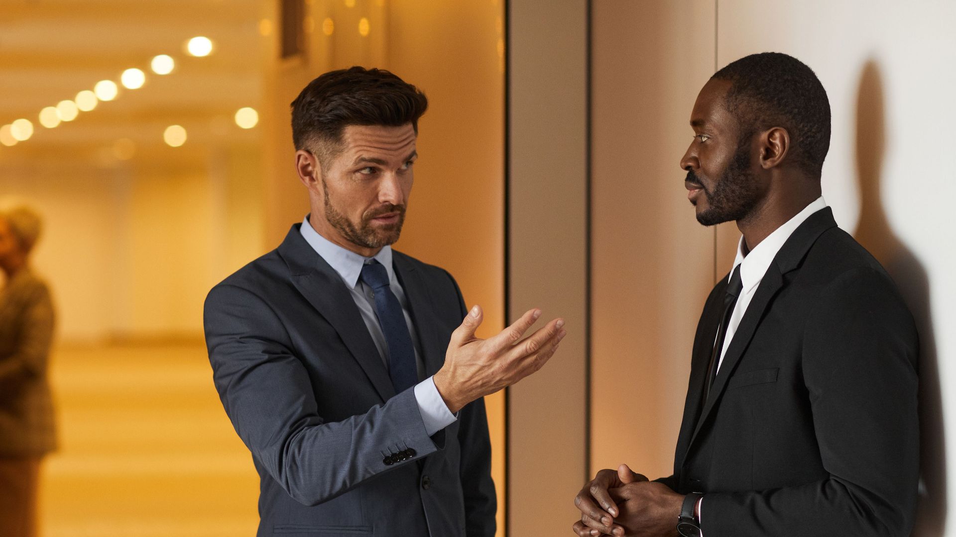 Two businessmen in formal attire engaged in a serious discussion in a modern office hallway.