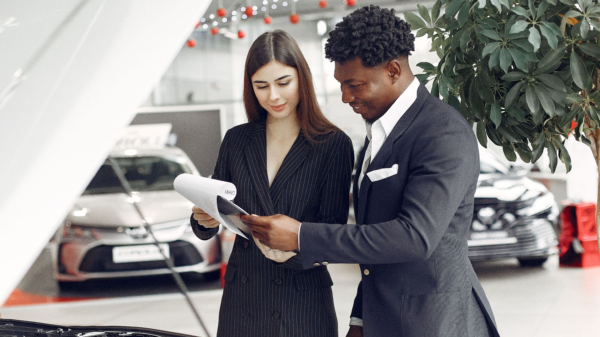 Confident smiling successful black businessman in classy formal suit standing near open motor hood in car showroom and discussing car characteristics with gorgeous female dealer