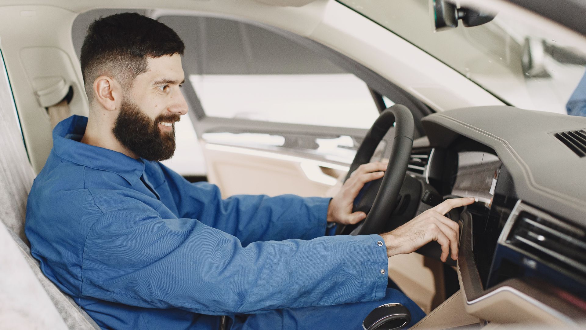 Mechanic in blue coveralls interacts with car dashboard, smiling and focused.