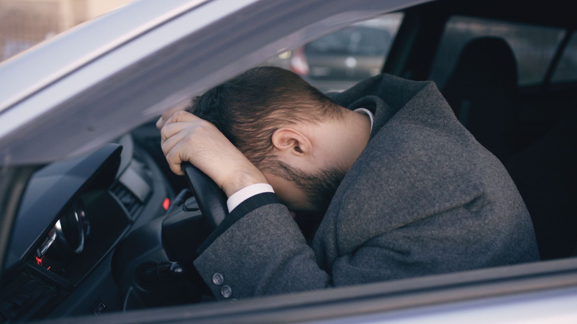 Man feeling overwhelmed and stressed, resting his head on a steering wheel inside a car.