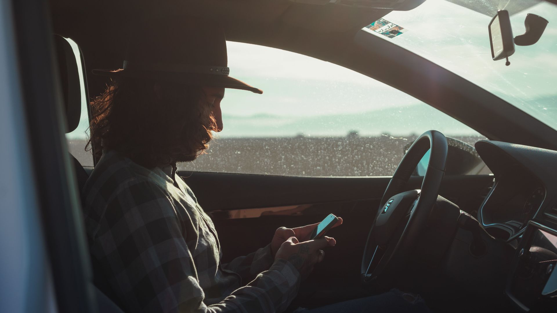 Profile of a man with a hat using a phone inside a car during the day.