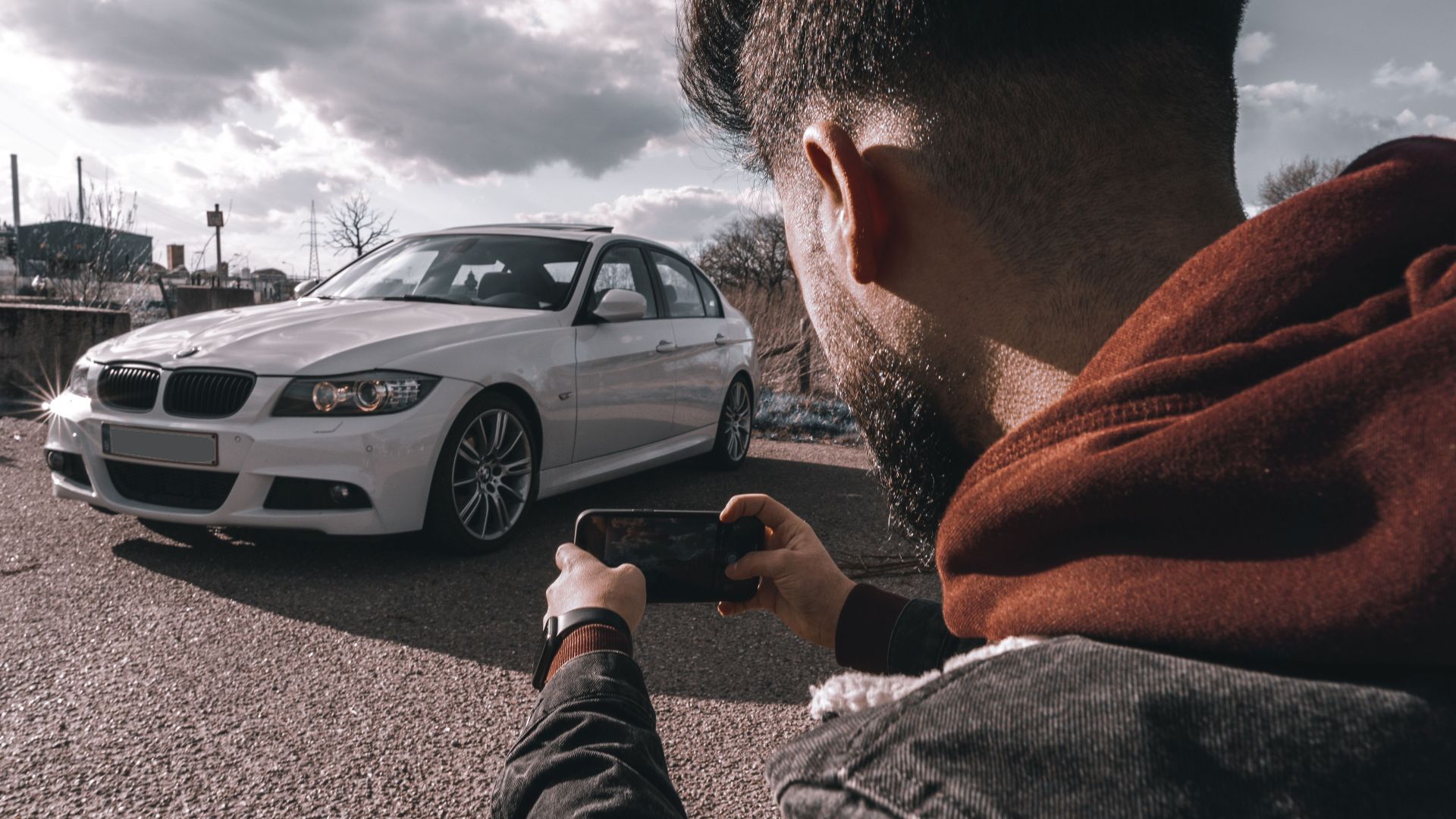 A man takes a picture of a white luxury car using a smartphone outdoors under a cloudy sky.