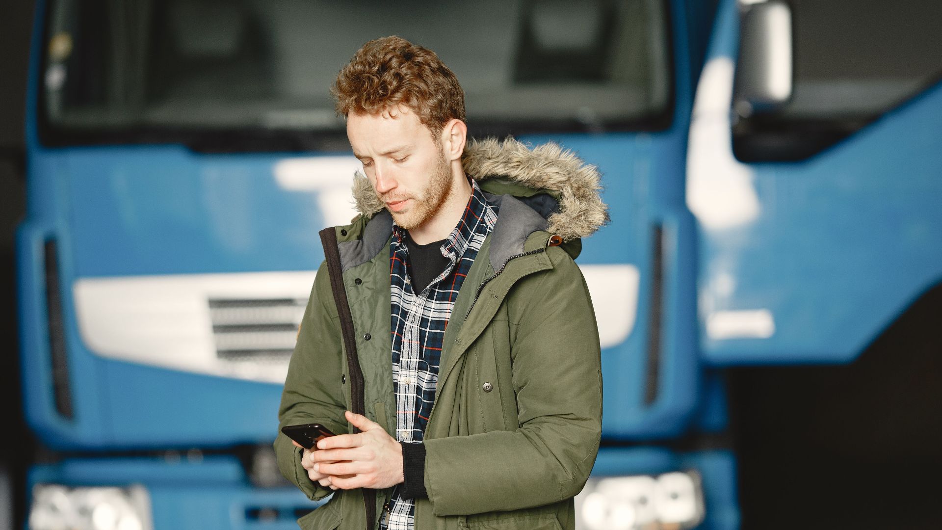 A man wearing a parka checks his mobile phone in front of a blue truck parked in a garage.