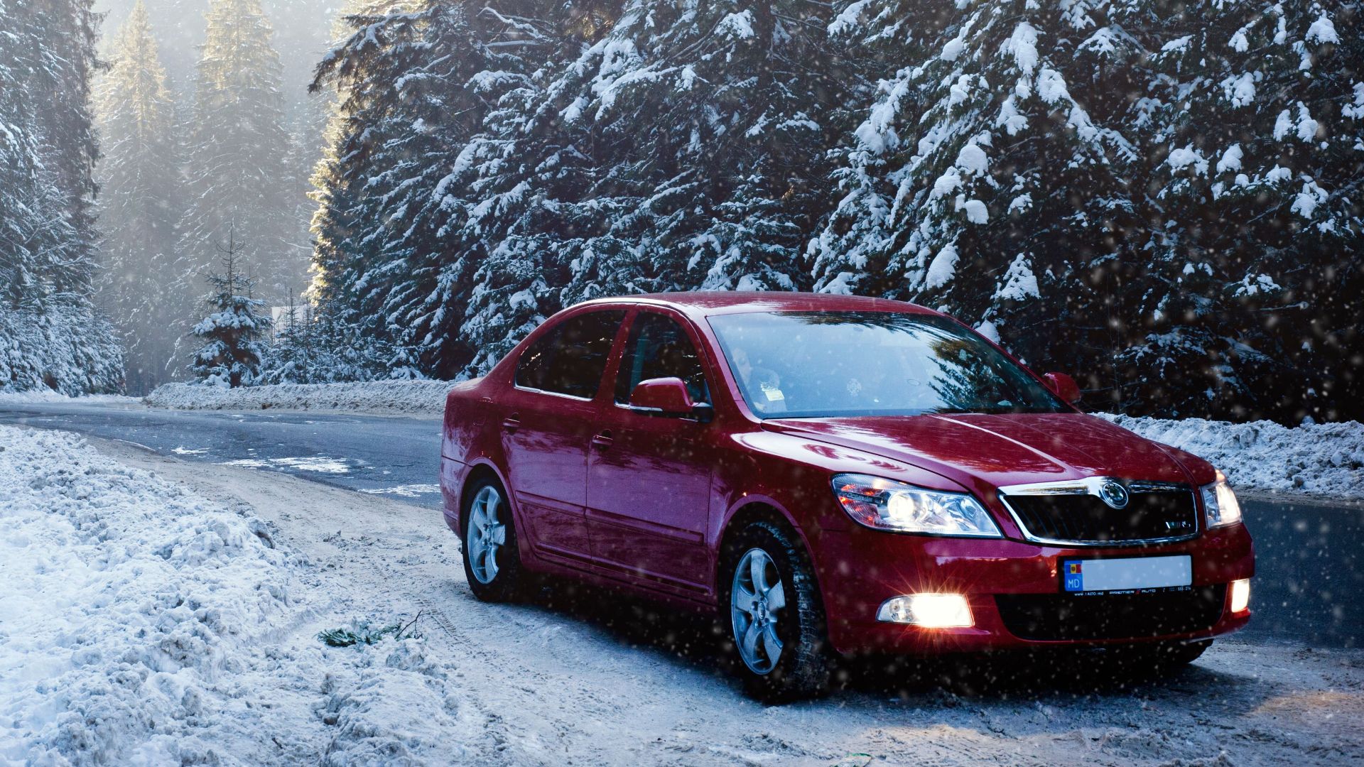 A red car drives along a snowy forest road in winter, surrounded by frosty trees.