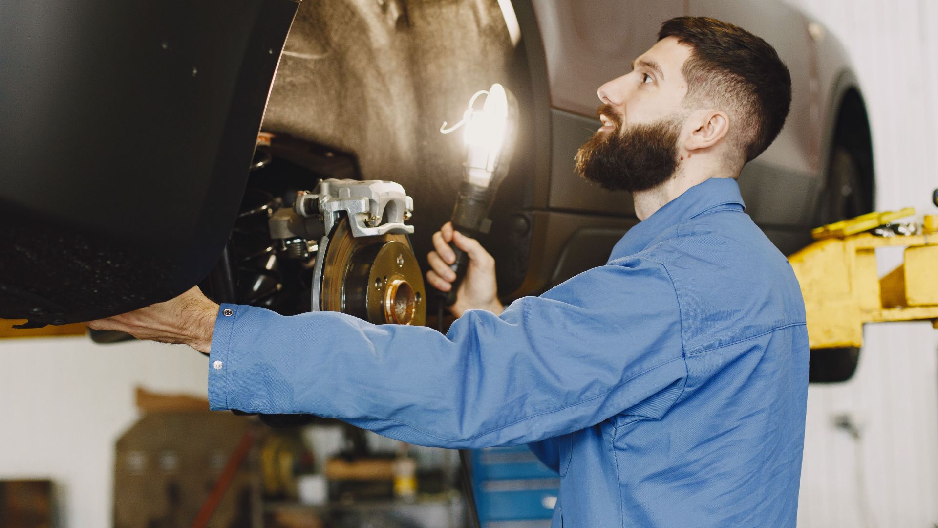 Professional mechanic in blue coveralls inspecting car brakes in a well-lit garage.