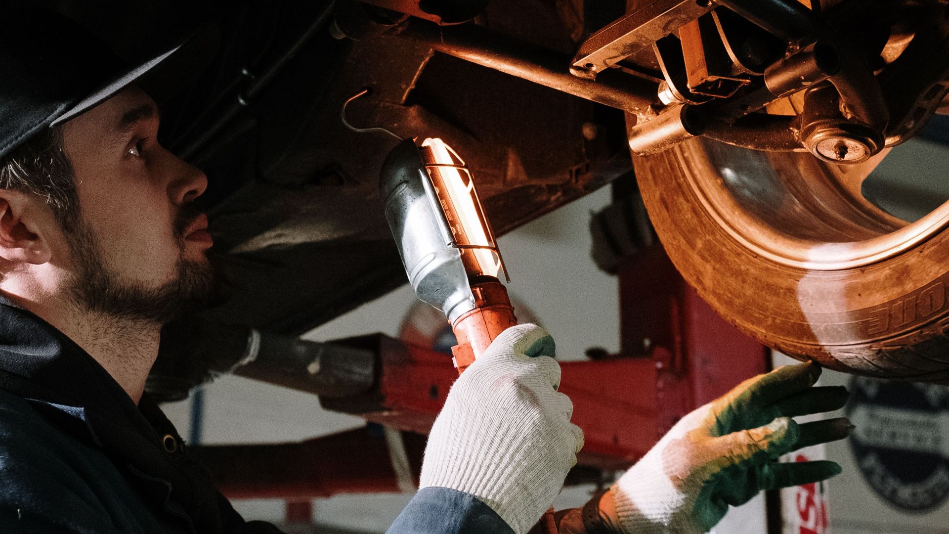 Mechanic uses a flashlight to inspect car's suspension in a garage setting.