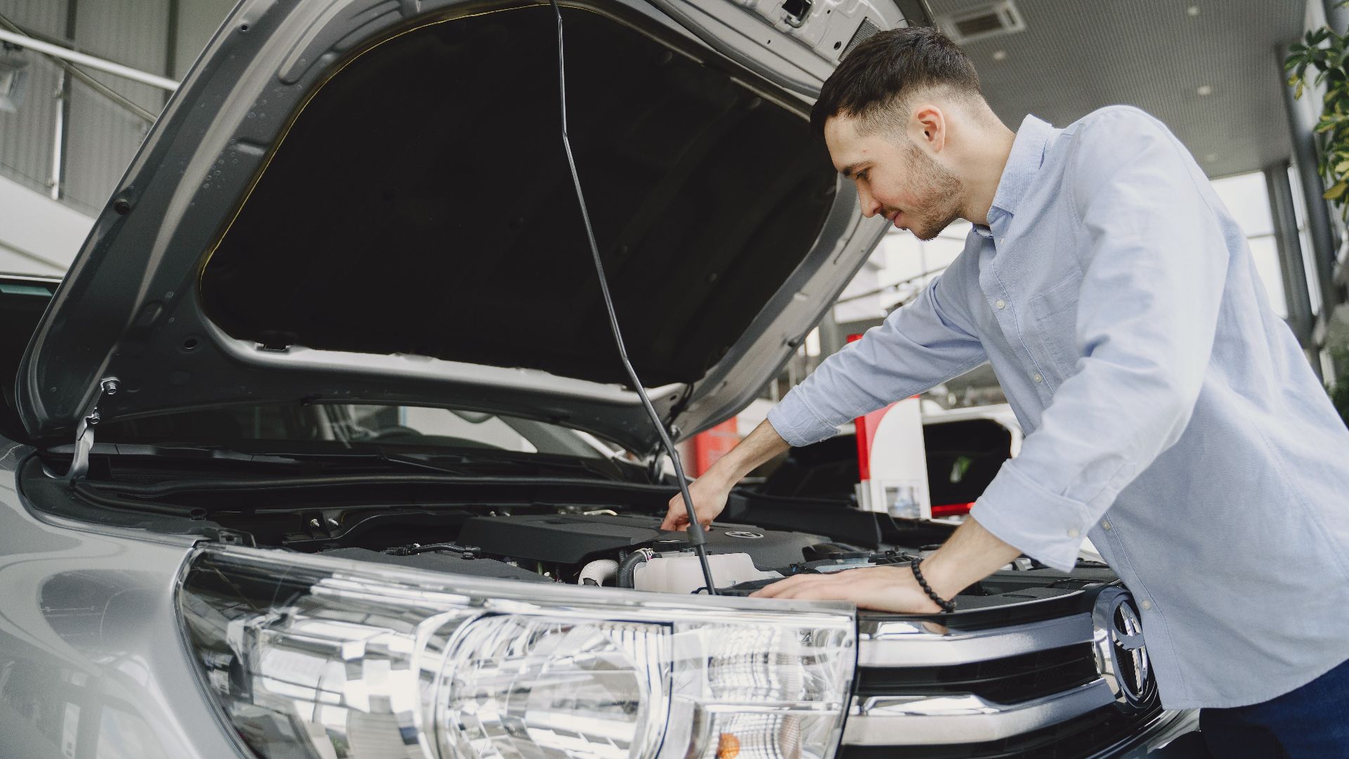 A man in a blue shirt examines a car's engine in a modern vehicle showroom.