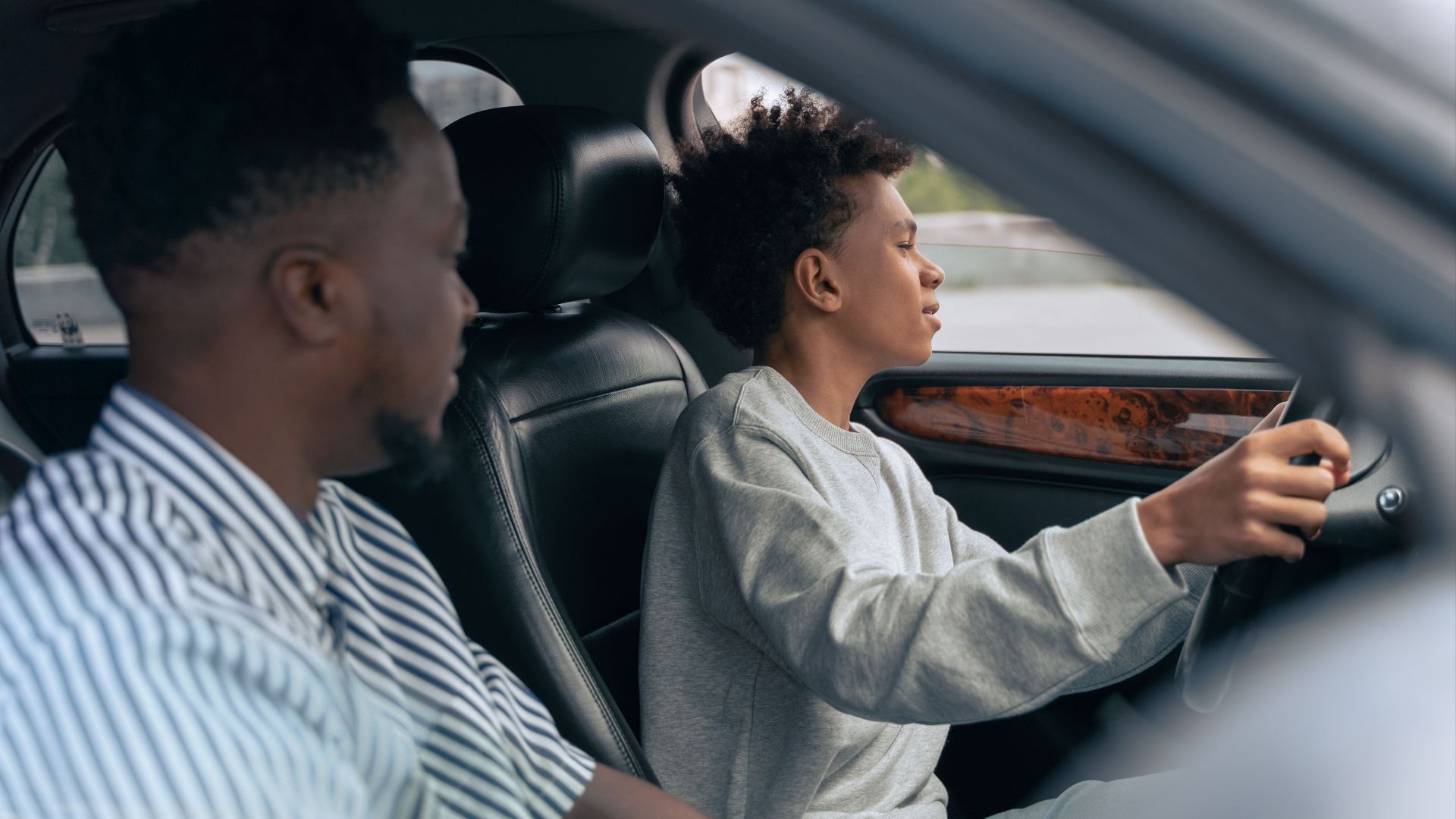 A father teaches his son to drive, sitting together in a car and discussing techniques. Side view indoors.