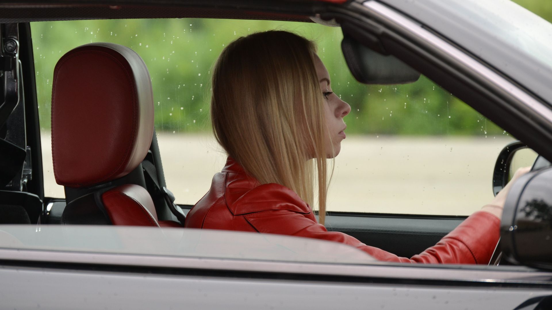 A young woman in a red convertible driving on a rainy day, wearing a red jacket.