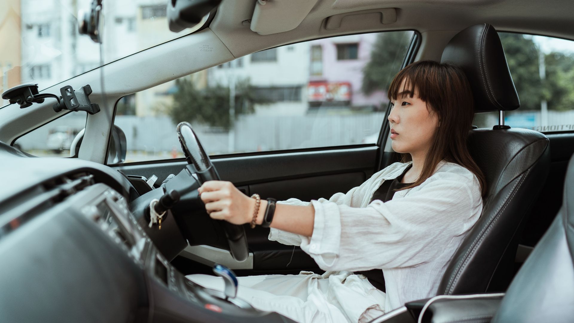 Side view unemotional Asian female in casual white clothes steering right hand drive car along urban streets in daytime
