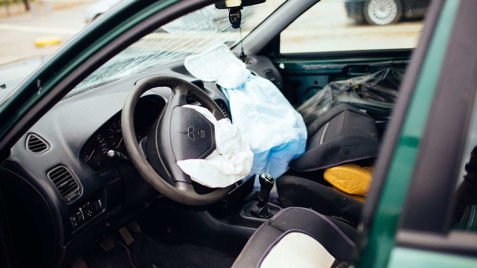 The car interior shows severe damage from a crash, with the airbag deployed and debris scattered. The accident took place on a busy urban street during daylight hours.