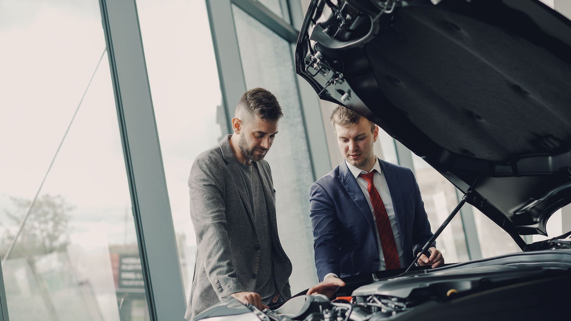 Two men inspecting a car's engine hood in a dealership showroom.
