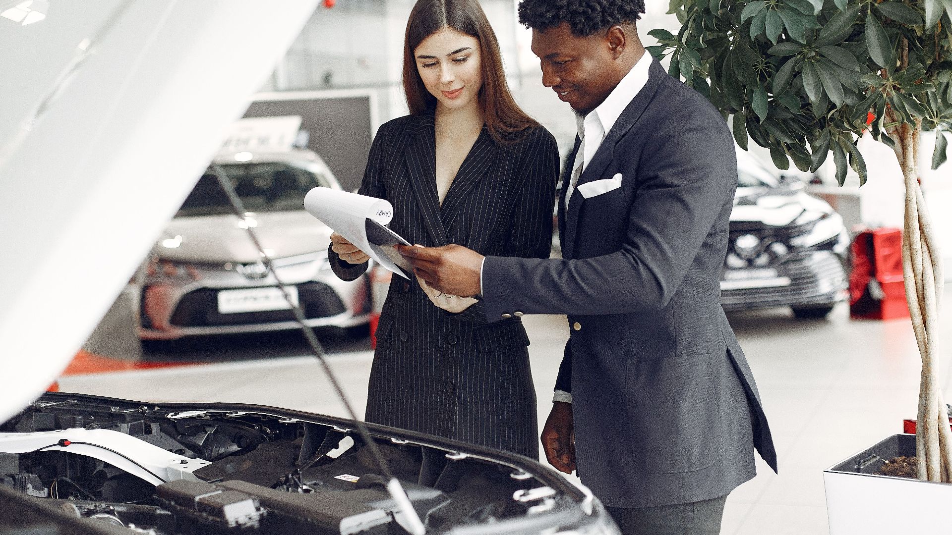 Confident smiling successful black businessman in classy formal suit standing near open motor hood in car showroom and discussing car characteristics with gorgeous female dealer