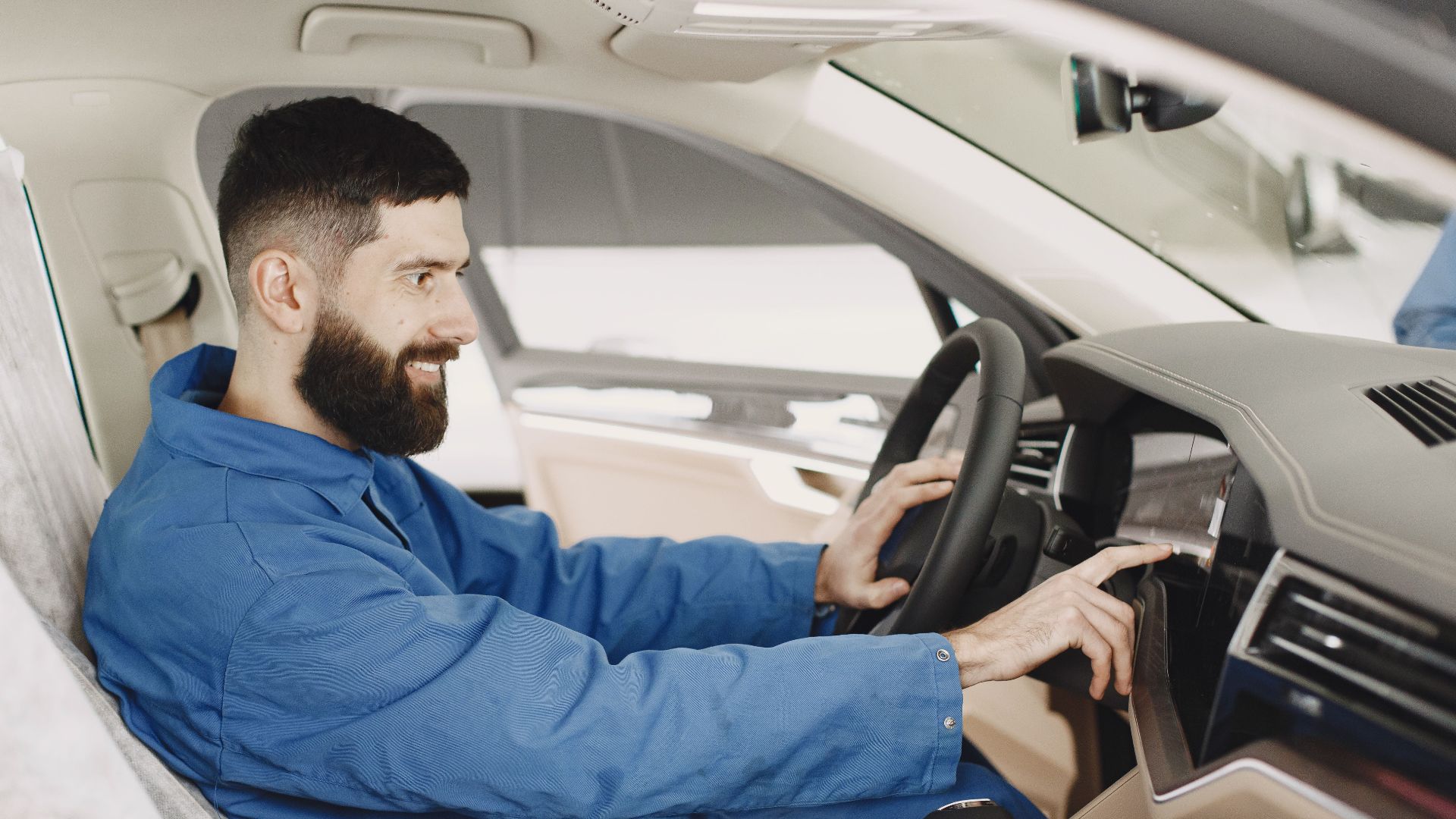 Mechanic in blue coveralls interacts with car dashboard, smiling and focused.