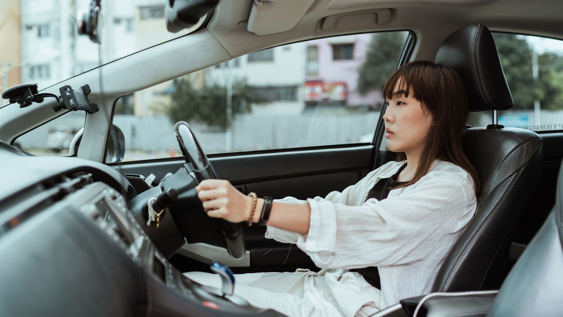 Side view unemotional Asian female in casual white clothes steering right hand drive car along urban streets in daytime