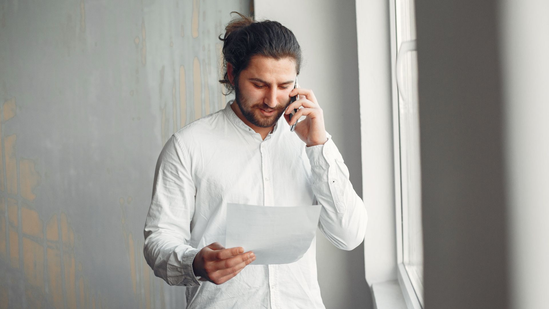 A young man in casual attire talks on the phone while holding a document indoors.