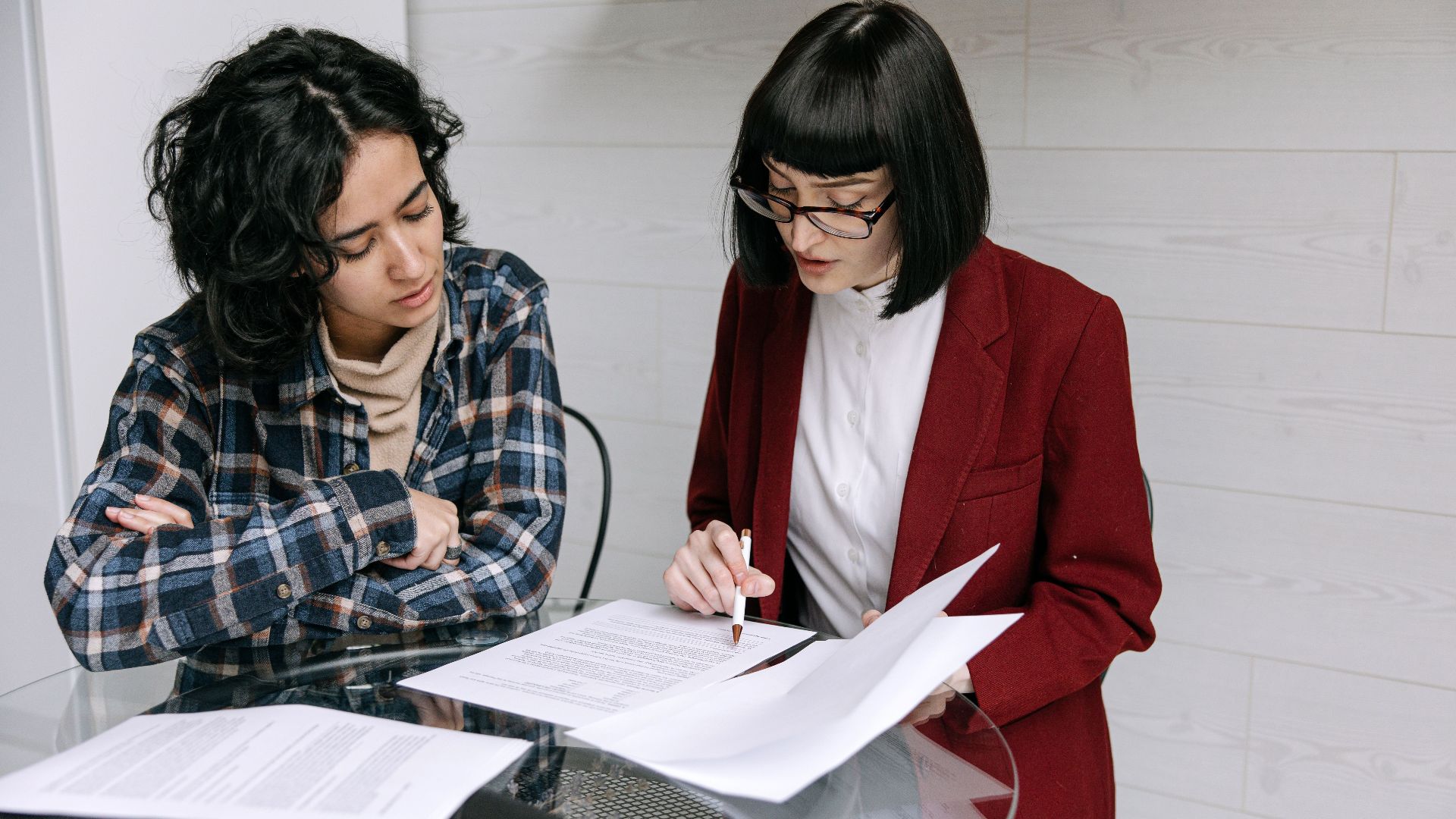 Two women collaborating at a glass table, reviewing business documents in a modern office setting.