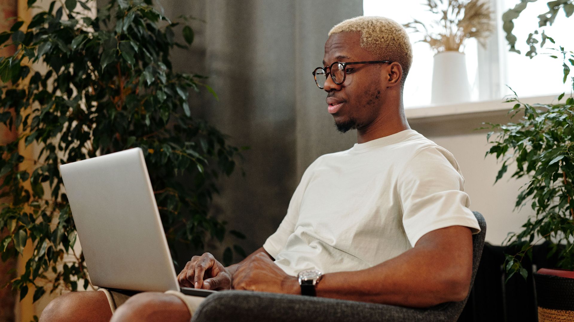 Focused young man with glasses working on a laptop in a cozy indoor space surrounded by plants.