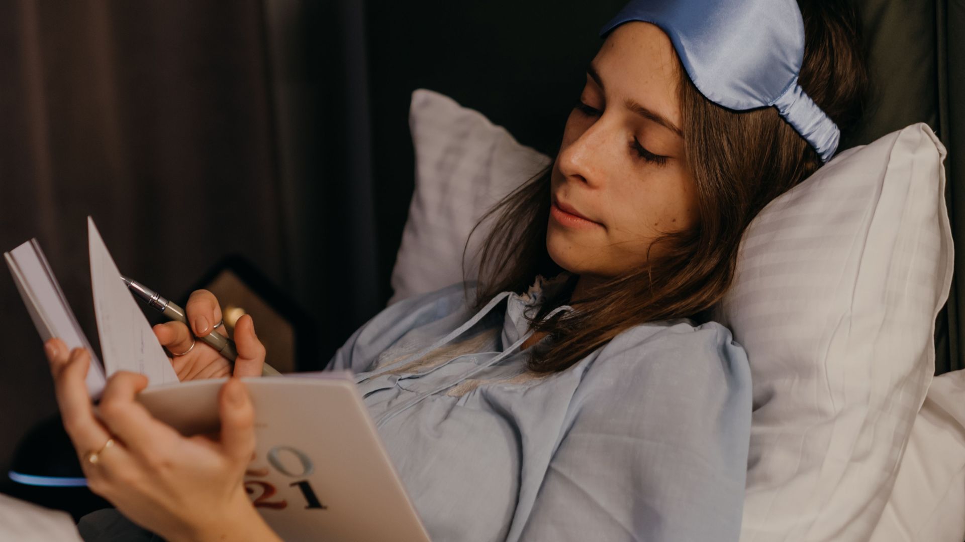 A young woman relaxing and writing in her journal while resting in bed with a sleep mask.