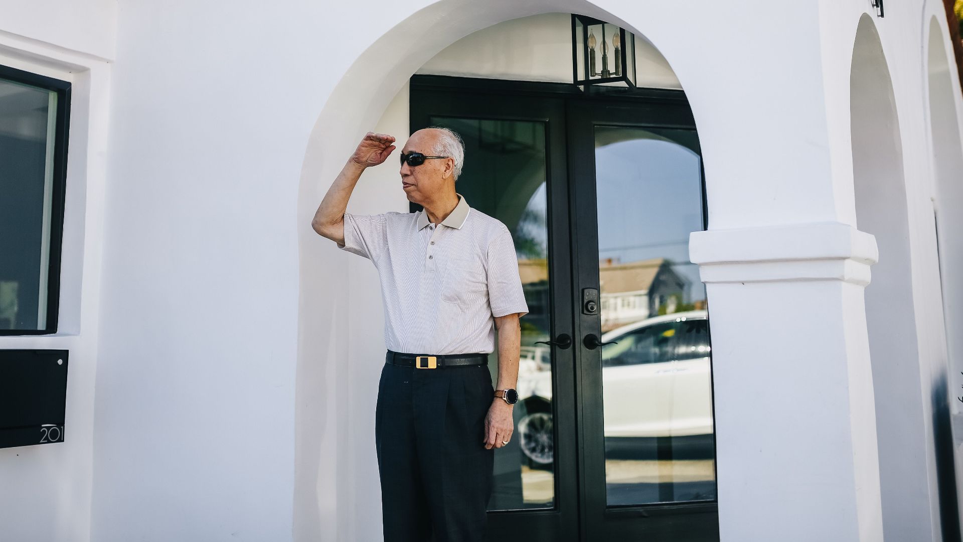 Elderly man in sunglasses standing outdoors by a white house with red-tiled roof.