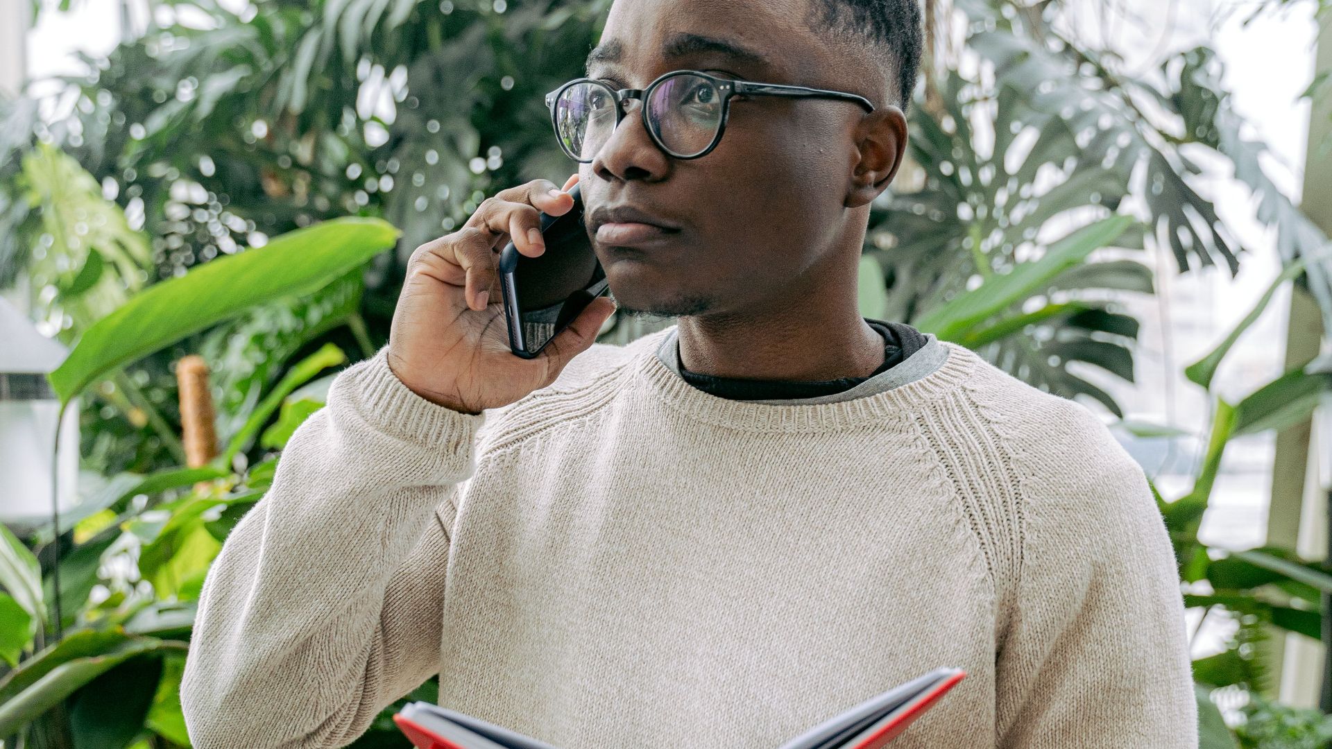 Adult man wearing eyeglasses, using smartphone and reading notebook indoors.