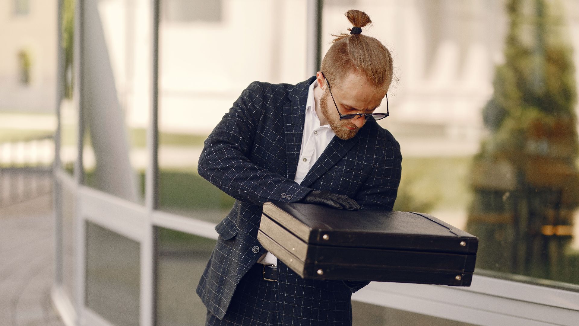 Businessman with a briefcase examines documents outside a modern building in the daytime.